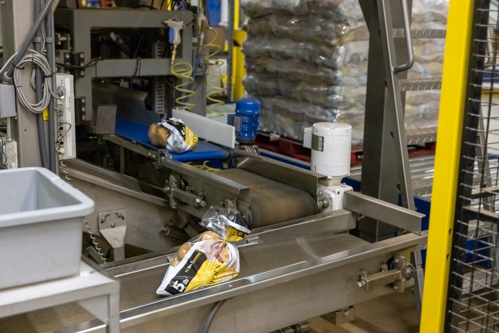 Snack bags on a metal conveyor in an automated packaging line at a factory, with gray machinery nearby.